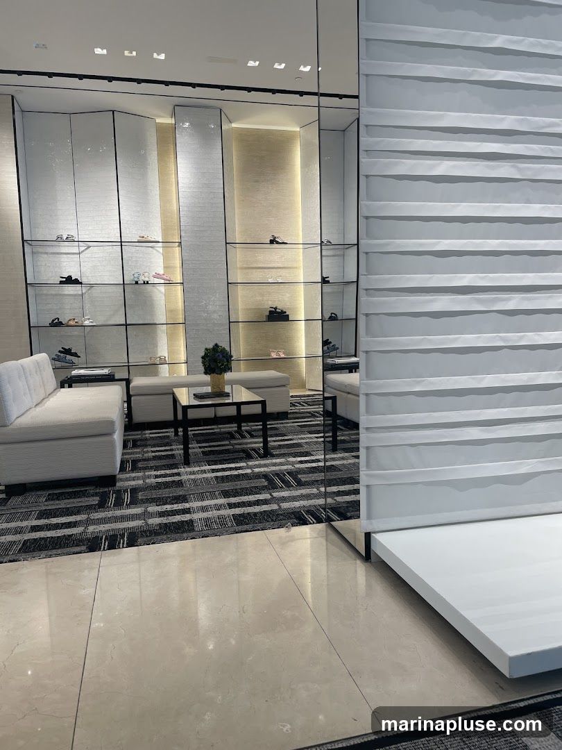 A well-lit interior of a high-end boutique, featuring a display of women's shoes on tiered shelves. The shelves are integrated into a modern, textured wall. In the foreground, a plush white sofa and a minimalist coffee table with a decorative plant create a seating area. The floor is a mix of polished marble and a patterned carpet. A large mirror reflects the display and seating area, adding to the spacious feel of the store.