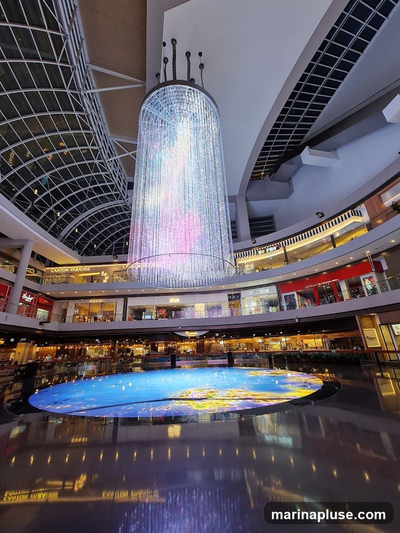An interior view of a modern shopping mall featuring a large, cylindrical chandelier made of numerous lights, suspended from the ceiling. Below the chandelier is a circular digital display on the floor, showing a vibrant, colorful image. The mall has multiple levels with shops and restaurants visible on the balconies surrounding the central atrium. The floor is highly reflective, mirroring the lights and architecture.