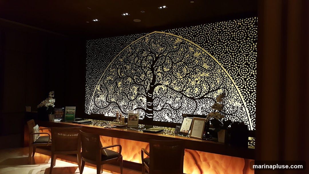 A reception desk with chairs in front of a large, ornate wall decoration featuring a stylized tree with intricate leaf patterns. The desk has several items on it, including brochures and framed documents. The wall behind the desk is illuminated from behind, highlighting the detailed cut-out design.