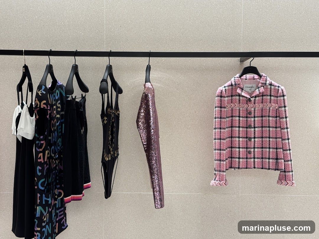 A black rack displays several pieces of Chanel clothing against a textured gray wall. From left to right, there is a white ruffled top over a black dress with colorful Chanel logos, a black skirt with striped trim, a black swimsuit with gold embellishments, a shimmering pink sequined jacket, and a pink and white plaid tweed jacket with a chevron pattern detail. The Chanel logo is visible on the label of the pink jacket.
