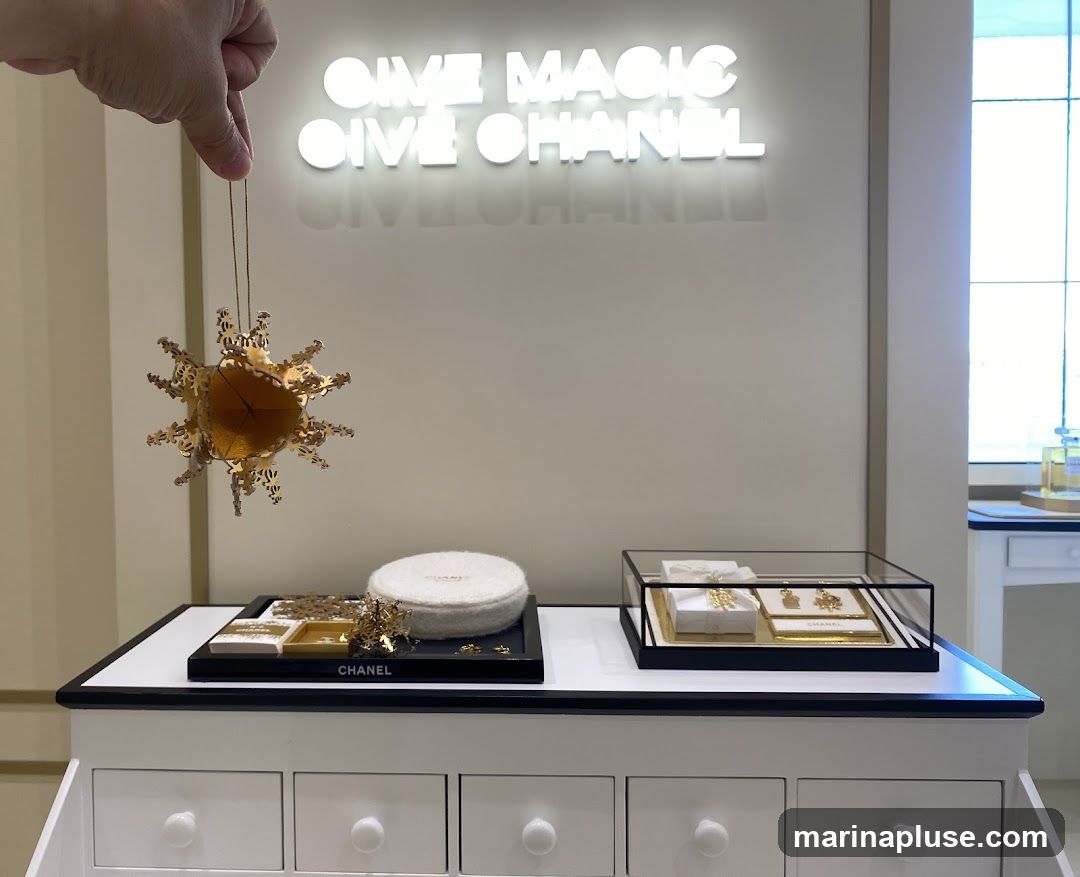A hand holds a golden, star-shaped ornament over a display of Chanel jewelry and cosmetics. The background features a neon sign that reads "GIVE MAGIC GIVE CHANEL".