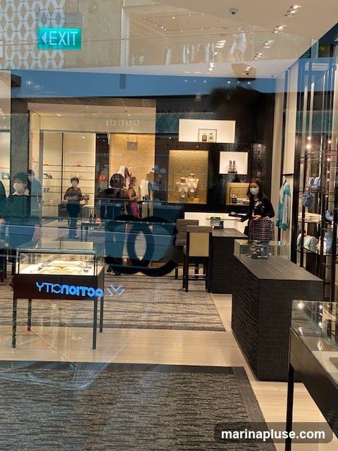 View through a glass storefront into a high-end retail store. The store features display cases with jewelry and accessories, mannequins dressed in clothing, and elegant interior design with dark wood accents and textured walls. Several customers and staff members, some wearing masks, are visible inside. An illuminated "EXIT" sign is visible above the entrance.