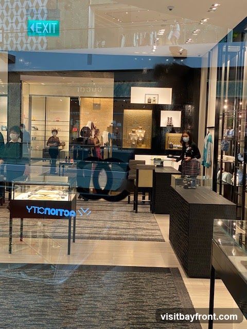 View through a glass storefront into a high-end retail store. The store features display cases with jewelry and accessories, mannequins dressed in clothing, and elegant interior design with dark wood accents and textured walls. Several customers and staff members, some wearing masks, are visible inside. An illuminated "EXIT" sign is visible above the entrance.