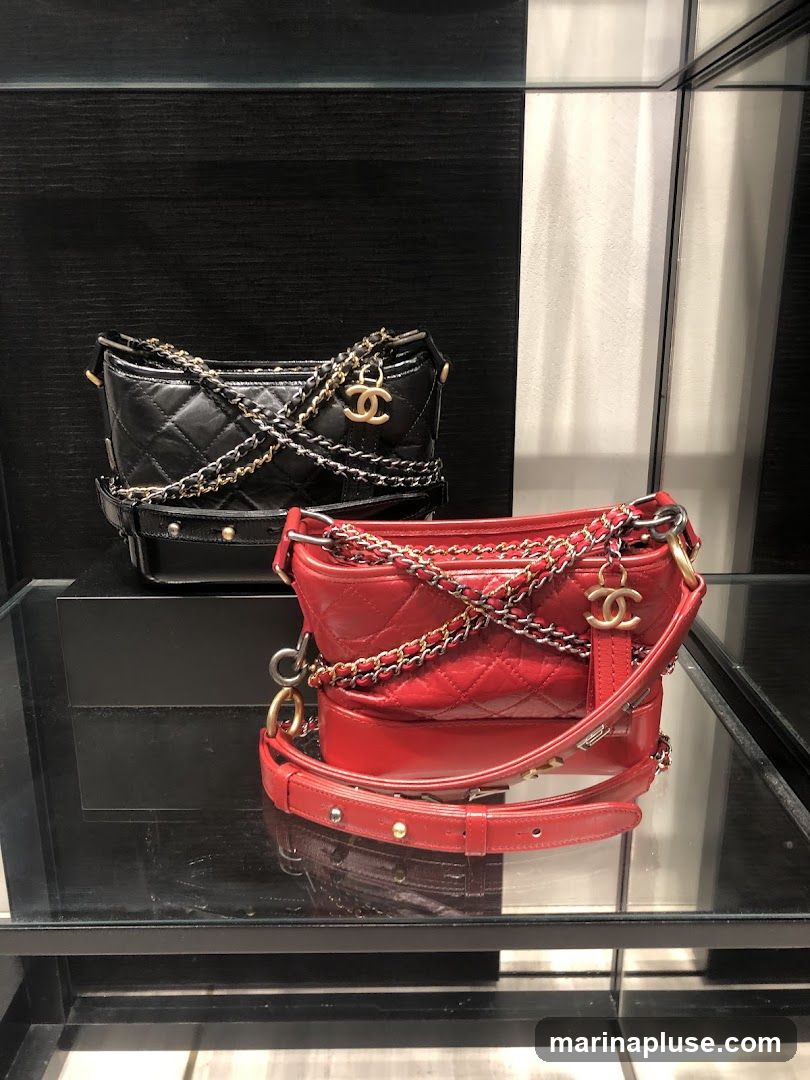 Two Chanel handbags, one black and one red, are displayed on a shelf. Both bags are quilted and feature multiple chain straps and a gold Chanel logo charm.