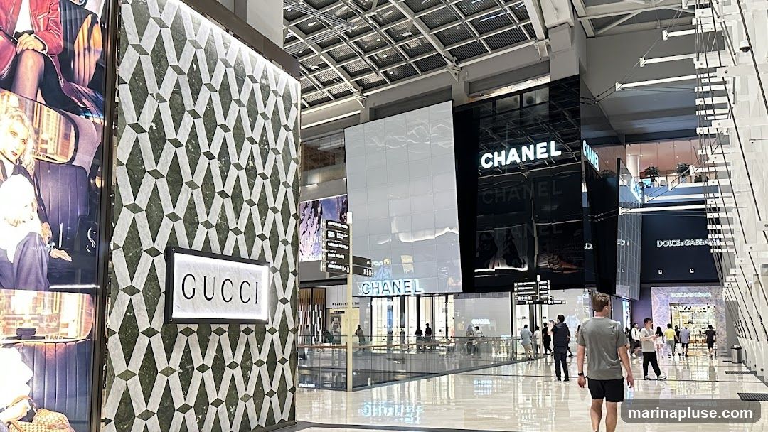 A wide shot of a modern, upscale shopping mall interior. On the left, a Gucci store facade is visible with its distinctive green and white geometric pattern and illuminated logo. Further down the mall, large illuminated signs for Chanel and Dolce & Gabbana are prominent. People are seen walking through the spacious, well-lit mall, with reflections on the polished floor. The architecture features a high ceiling with exposed structural elements and lighting.
