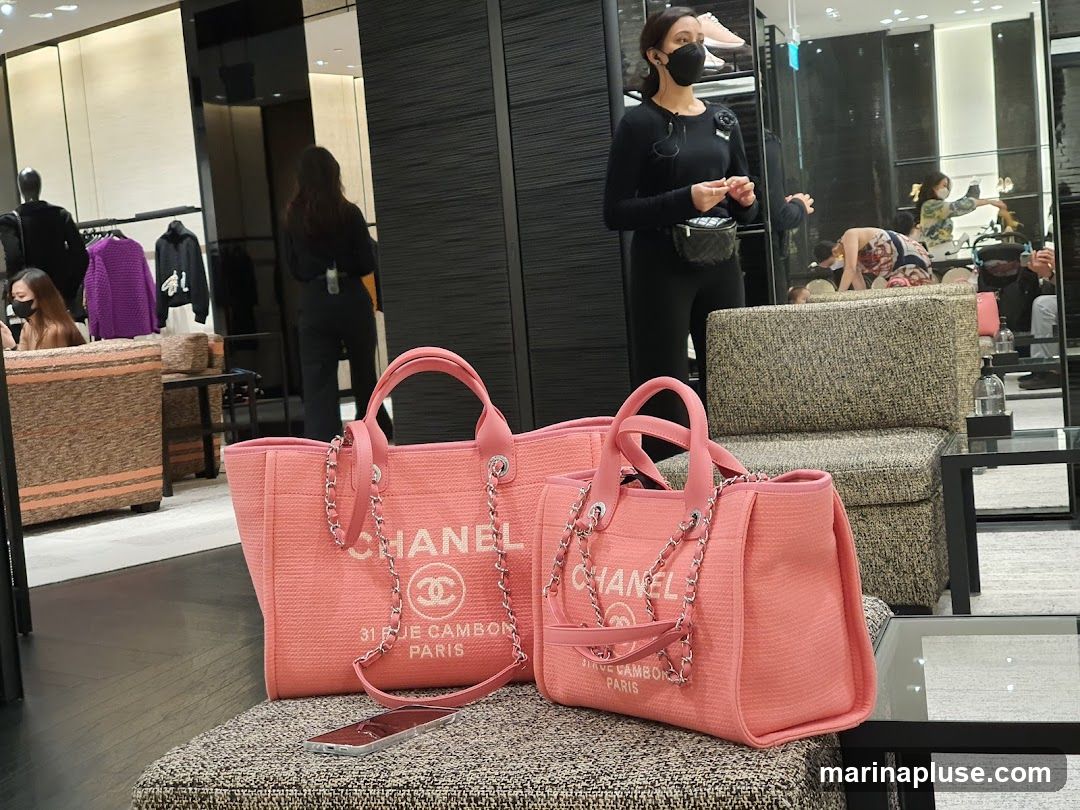 Two pink Chanel tote bags are displayed on a textured ottoman in a luxury retail store. The bags feature the Chanel logo and "31 RUE CAMBON PARIS" text. In the background, a sales associate in black attire and a face mask is visible, along with other customers and merchandise on display.