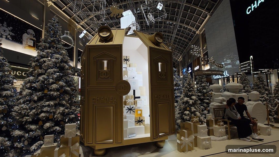 A festive winter display featuring a large, golden Chanel-branded house structure filled with gift boxes and perfume bottles. Snow-covered Christmas trees and decorative presents surround the display. In the background, other luxury brand storefronts are visible, including Gucci. Two people are seated on a bench in front of a large perfume bottle sculpture.