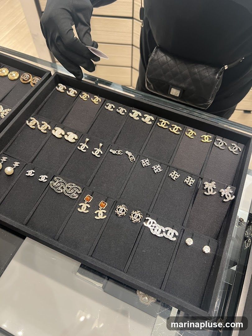 A display case filled with various Chanel earrings. The earrings are arranged on black velvet stands within a black tray. A person wearing black gloves and a black quilted fanny pack is visible in the background, holding a small white card.