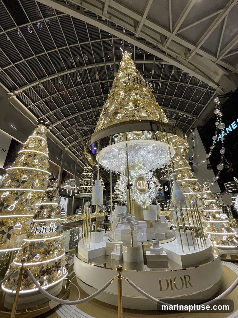 A festive indoor display featuring multiple illuminated Christmas trees decorated with Dior branding and products. The central display is a multi-tiered platform showcasing Dior perfumes and gift boxes, adorned with white sculptures and golden accents. The overall scene is illuminated with warm, festive lighting, creating a luxurious holiday atmosphere.