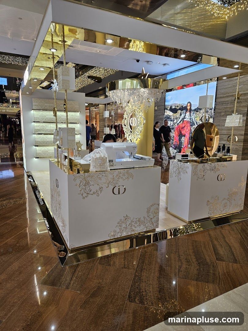 A white and gold kiosk with Dior branding is displayed in a shopping mall. The kiosk features shelves stocked with products, a large screen displaying an advertisement, and decorative elements. Several people are visible browsing the merchandise.