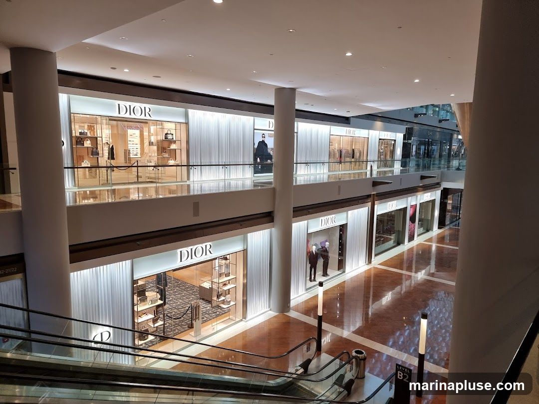 A wide shot of a multi-level shopping mall interior, focusing on several Dior storefronts. The stores are illuminated and display handbags, clothing, and accessories. Escalators are visible in the foreground, and the mall's polished floors reflect the lights.