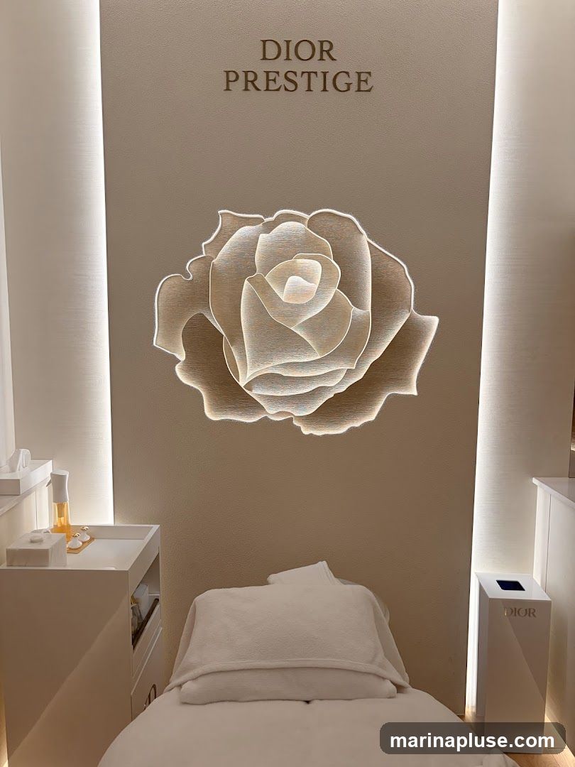 A treatment room with a massage bed covered in white linens. Above the bed, a large, illuminated rose sculpture is mounted on a textured wall. The words "DIOR PRESTIGE" are displayed above the rose. To the left, a white trolley holds skincare products, including a bottle of serum and small jars. To the right, a white pedestal with the Dior logo is visible.