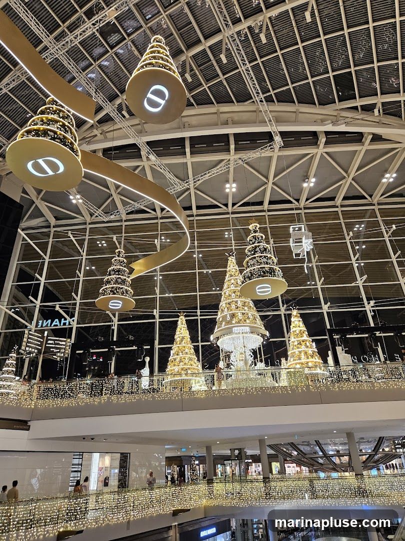 A festive indoor scene with multiple levels of a mall decorated for Christmas. Large, illuminated Christmas trees are displayed on the upper levels, some adorned with lights and ornaments. Hanging from the ceiling are large, circular ornaments with a logo, possibly from a brand like Dior. A golden ribbon-like structure curves through the space, also illuminated. The railings of the upper floors are draped with strings of fairy lights, creating a warm and inviting ambiance. Several shops are visible on the lower levels, with some shoppers browsing. The overall impression is one of a grand, festive shopping environment.