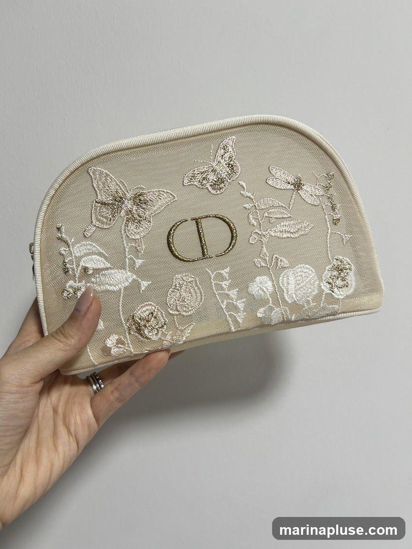 A hand holds a beige Dior cosmetic pouch with intricate embroidery of butterflies, dragonflies, and floral motifs. The Dior CD logo is prominently displayed in the center. The pouch has a mesh overlay and a white trim.