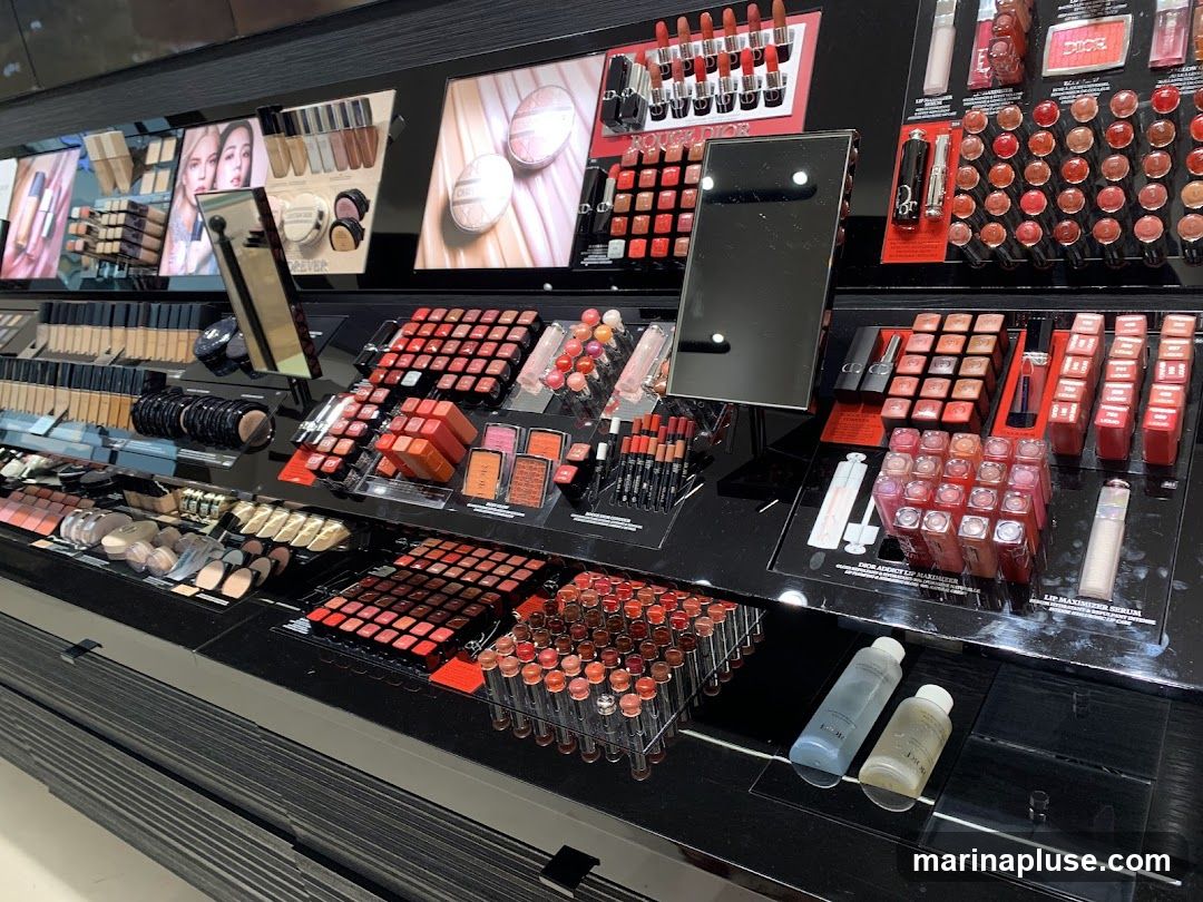 A display of various makeup products, including lipsticks, foundations, blushes, and lip glosses, arranged on shelves. Some products have visible branding, such as Dior. There are also promotional images of models and product swatches.