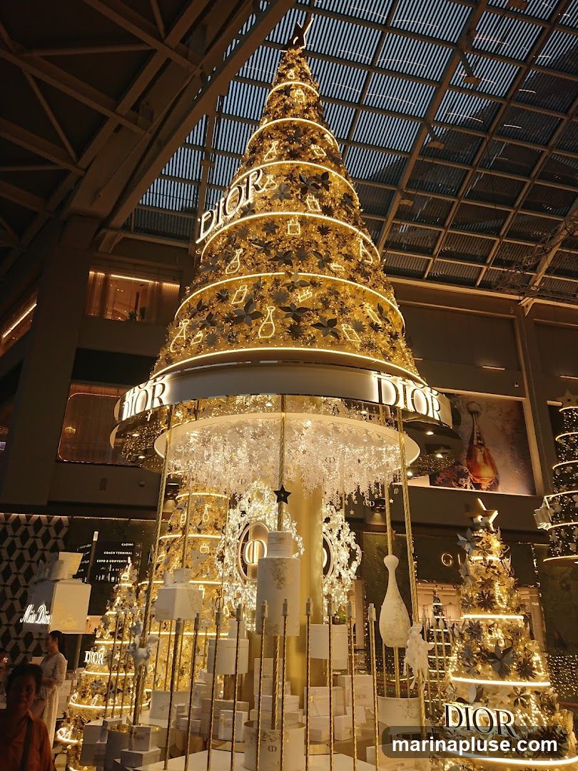 A large, elaborately decorated Christmas tree with Dior branding is displayed indoors, likely in a shopping mall. The tree is adorned with lights, ornaments shaped like perfume bottles and flowers, and prominent Dior logos. Surrounding the main tree are smaller decorative trees and displays of Dior products, all in a festive gold and white color scheme.