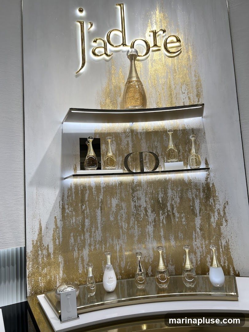 A display featuring various bottles of Dior J'adore perfume on shelves. The background is white with gold abstract splatters. The J'adore logo is prominently displayed in gold lettering at the top. A small card with the J'adore logo and "Dior" is visible on the bottom shelf.