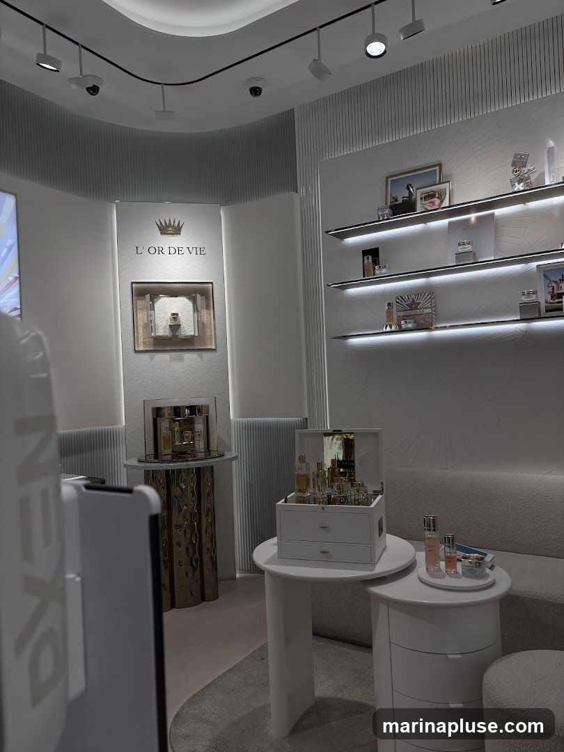 A well-lit interior of a cosmetics store featuring various products displayed on shelves and tables. The brand "L'OR DE VIE" is visible on a wall display with a crown logo. Products include perfumes, skincare items, and makeup palettes. A white sofa is visible in the background.
