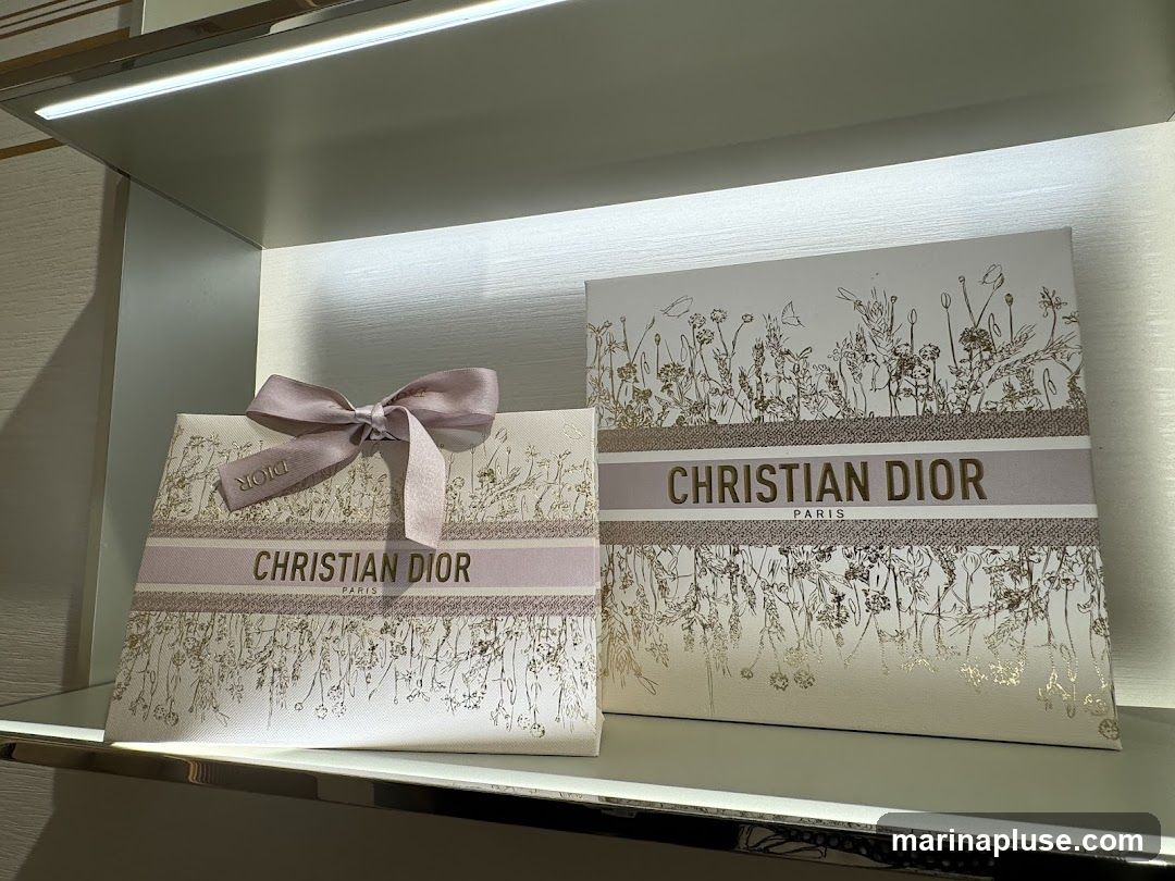 Two gift boxes from Christian Dior are displayed on a shelf. The boxes are white with a delicate floral pattern in gold and pink. A band across the center of each box features the "CHRISTIAN DIOR PARIS" logo. One box is tied with a light purple ribbon.