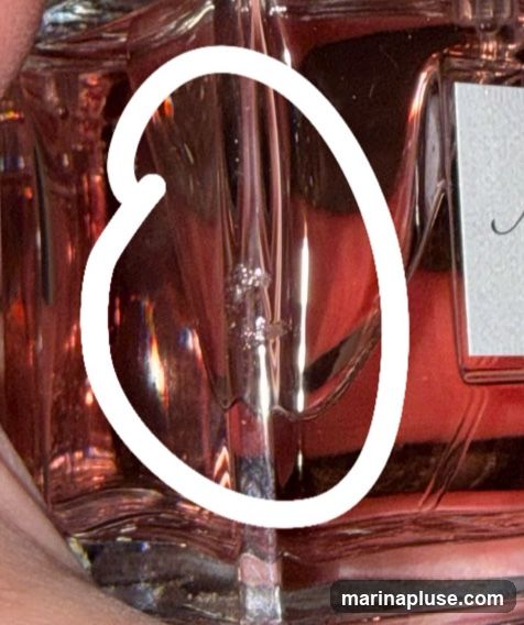 A close-up shot focuses on the glass bottle of a perfume, highlighting a section with small air bubbles trapped within the glass. The liquid inside the bottle appears to be a rosy or reddish hue. A white label with cursive text is partially visible on the right side of the bottle. The lighting creates reflections and highlights on the glass surface.