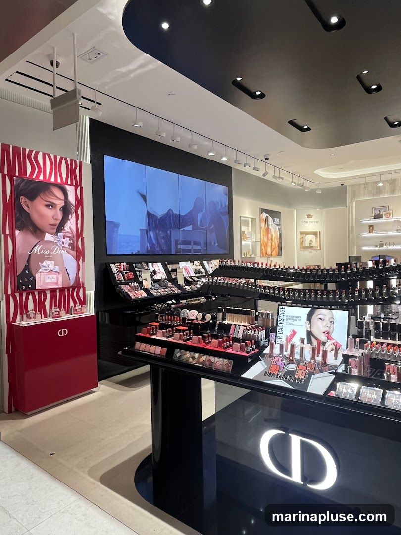 A well-lit interior view of a Dior cosmetics store. A large screen displays abstract imagery. Various makeup products, including lipsticks, eyeshadow palettes, and foundations, are neatly arranged on display shelves and tables. A prominent "Miss Dior" advertisement featuring a woman is visible on the left. The Dior logo is displayed on a black counter in the foreground.