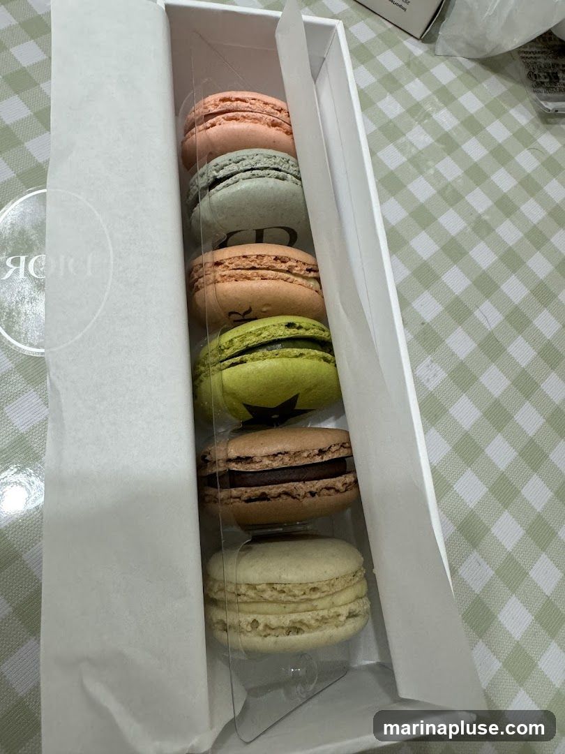 A white box containing six colorful macarons. The macarons are arranged in two columns of three. The colors include pink, light blue, orange, green, brown, and cream. The box has a white paper lining and a clear plastic insert to hold the macarons. The Dior logo is visible on the paper lining.
