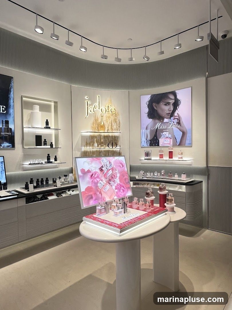 A well-lit interior display of Dior perfumes. A large screen shows an advertisement for Miss Dior perfume featuring Natalie Portman. In the foreground, a display table showcases various Miss Dior perfume bottles. Shelves in the background are stocked with different Dior fragrances and products, including Sauvage and J'adore. The overall aesthetic is clean, modern, and luxurious.