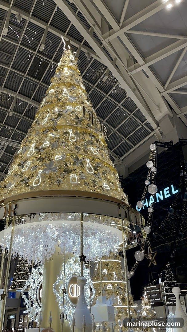 A large, elaborately decorated Christmas tree with Dior branding is prominently displayed indoors. The tree is adorned with lights and festive ornaments. In the background, a Chanel logo is visible, suggesting a luxury shopping environment. The overall scene is festive and opulent, likely part of a holiday display in a high-end retail space.