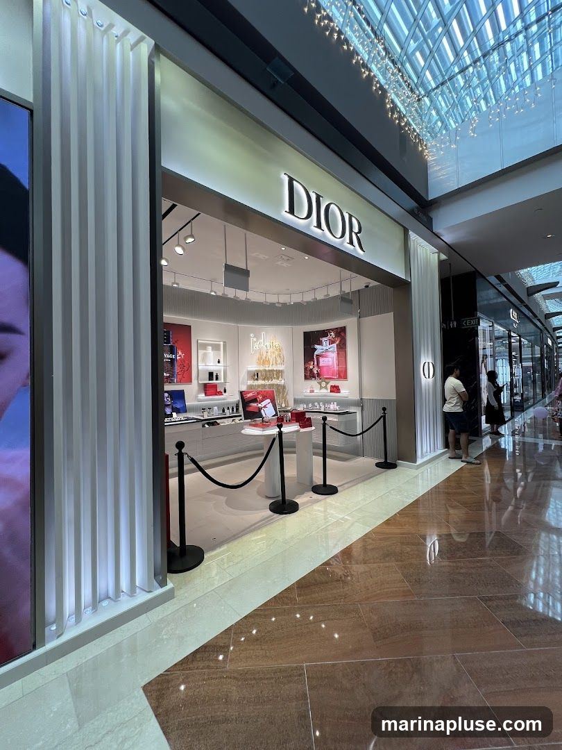 The exterior of a Dior store with the brand name prominently displayed above the entrance. The store features a clean, modern design with illuminated signage. Inside, various Dior beauty products are displayed on shelves and counters. The entrance is cordoned off with velvet ropes.