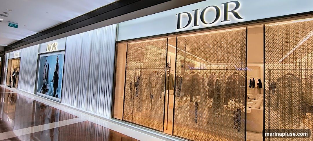 A wide shot of a Dior store's exterior in a mall. The facade features the prominent "DIOR" logo in illuminated lettering above a large display window. The window is covered by a decorative, woven-like screen, through which clothing items on racks are visible. To the left, another section of the store front displays a large advertisement featuring two mannequins. The store has a clean, modern aesthetic with white pleated drapes on the left side of the entrance.