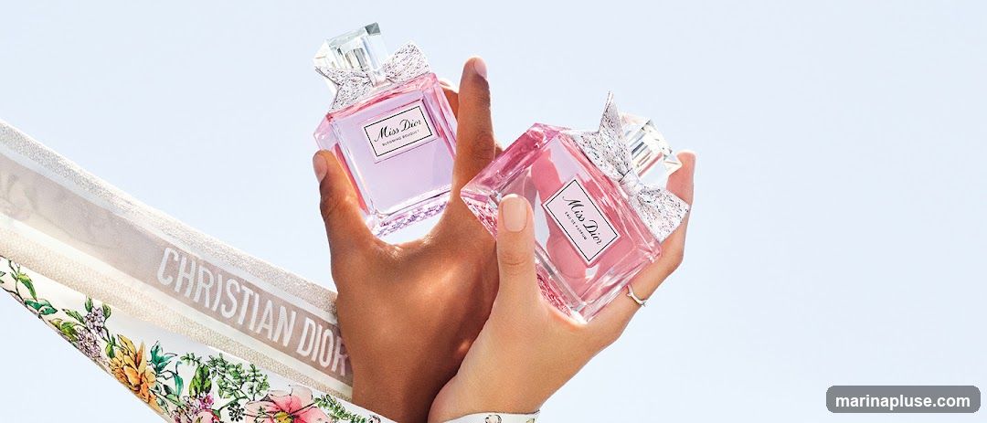Two hands hold up two bottles of Miss Dior perfume against a light blue sky. The bottles are pink and have a bow detail on the cap. A fabric with the Christian Dior logo and floral print is visible in the foreground.