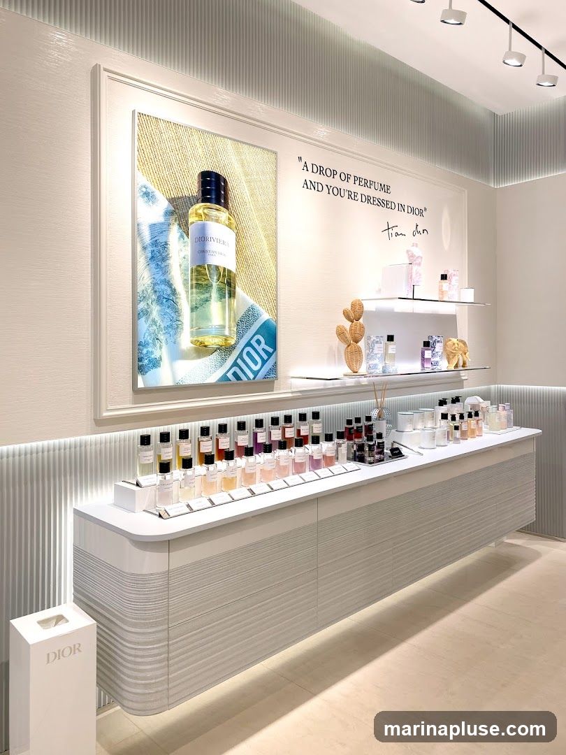 A well-lit display of Dior perfumes in a store. A large framed advertisement featuring a bottle of Dioriviera perfume is on the left. Text on the wall reads "A DROP OF PERFUME AND YOU'RE DRESSED IN DIOR" with a signature. A long counter displays numerous perfume bottles of various sizes and colors, along with some decorative items and diffusers. A white Dior branded bin is on the left.