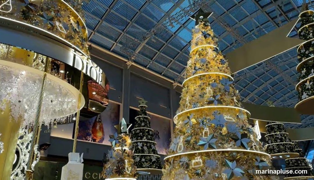A festive indoor scene featuring multiple elaborately decorated Christmas trees. The trees are adorned with gold and blue ornaments, lights, and star toppers. The background shows a modern ceiling structure and hints of store branding, including "DIOR" and "GUCCI".