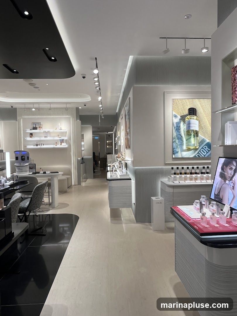 The interior of a well-lit Dior beauty store. Displays of perfumes, makeup, and skincare products are arranged on shelves and counters. A large advertisement featuring a model and Dior products is visible on the right wall. The store has a modern and luxurious design with white and grey tones.
