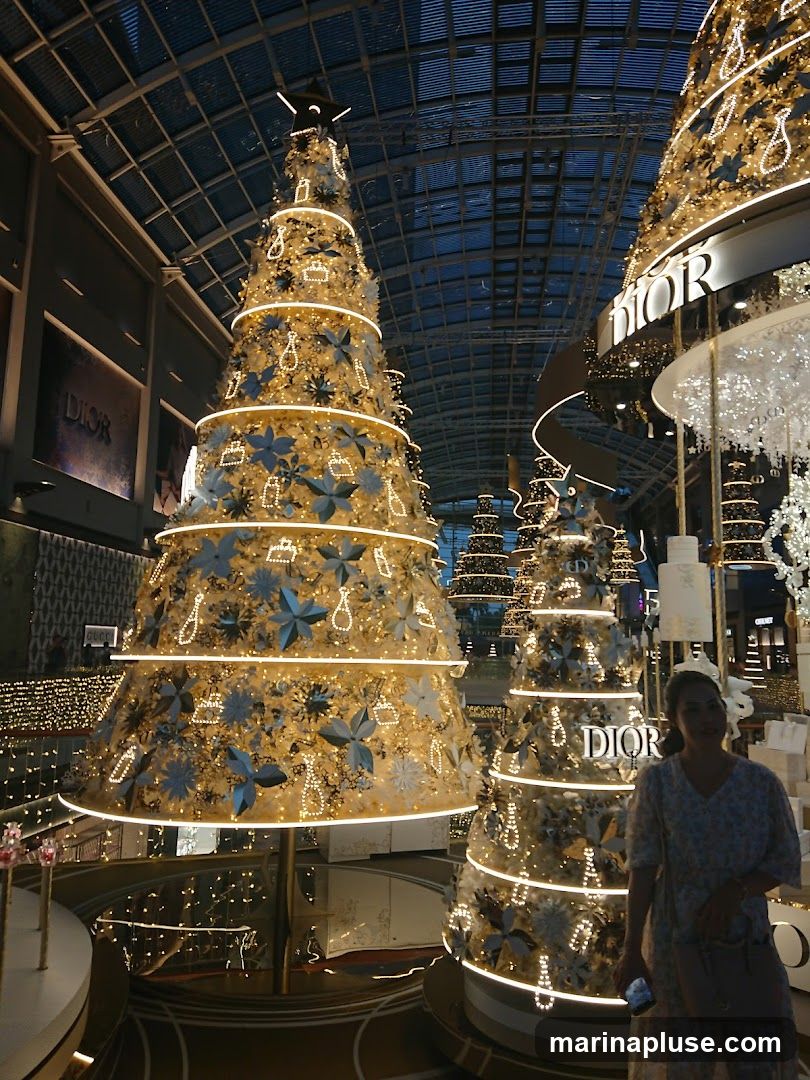 A festive display of large, illuminated Christmas trees decorated with ornaments and lights. The Dior logo is prominently displayed on some of the trees and on the surrounding structures, suggesting a holiday promotion or store decoration. A woman stands in the foreground, looking towards the trees. The scene appears to be indoors, possibly in a shopping mall or department store, with a glass ceiling visible above.