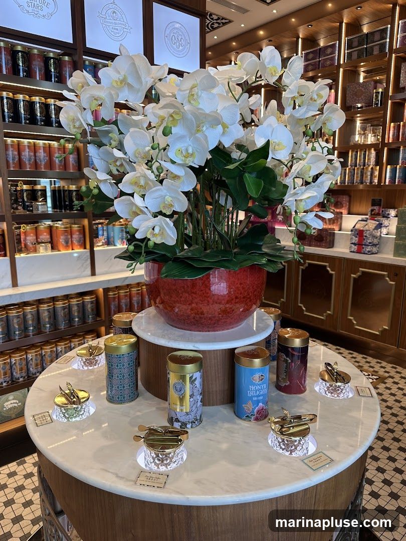 A display of decorative tins and small serving dishes on a marble-topped table, with a large arrangement of white orchids as a centerpiece. Shelves stocked with numerous tins are visible in the background.