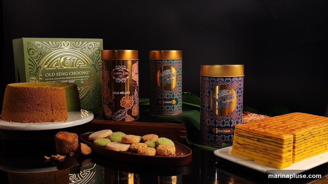A still life arrangement featuring a green chiffon cake, a slice of layered cake, assorted cookies, and three decorative tins. The tins are labeled with "Old Seng Choong," "Colonial," and "Kampong Spirit" with flavors like "Gula Melaka" and "Pandan." A green box with gold lettering also reads "Old Seng Choong."