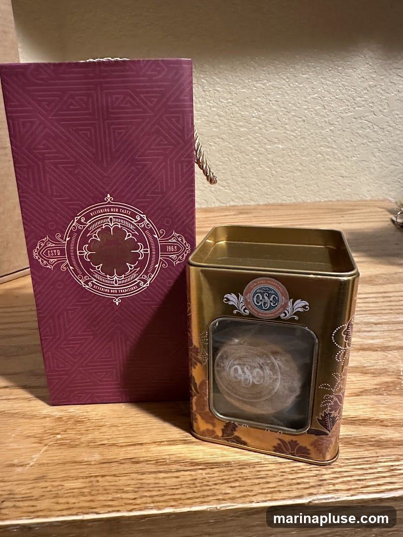 A tall, maroon gift box with a patterned design and gold accents stands next to a gold tin. The tin has a window revealing a round, light-brown item inside, also with a patterned design.