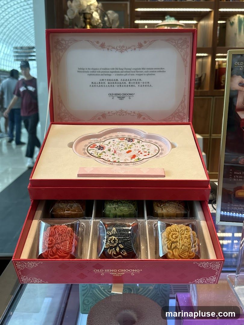 A red gift box is open, revealing a tray and a selection of mooncakes. The lid of the box has text describing the mooncakes and the brand "OLD SENG CHOONG". Below the tray are six mooncakes in individual packaging, arranged in two rows of three. The box has decorative patterns. To the right, a menu board is partially visible with prices for "Black T" and "Lotus Paste".