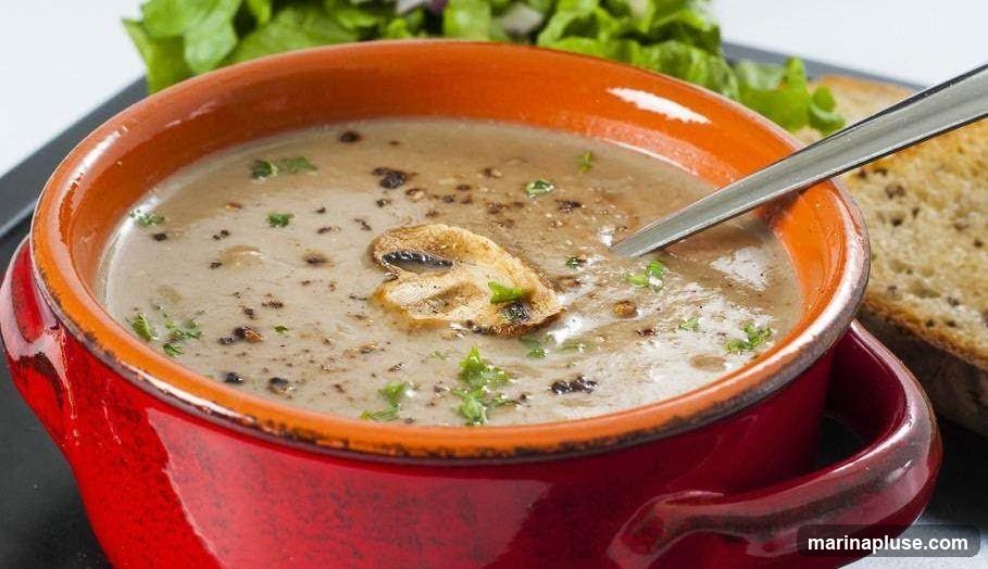 A close-up shot of a bowl of creamy mushroom soup, garnished with fresh parsley and a slice of mushroom. A piece of toasted bread and a small salad are visible in the background.
