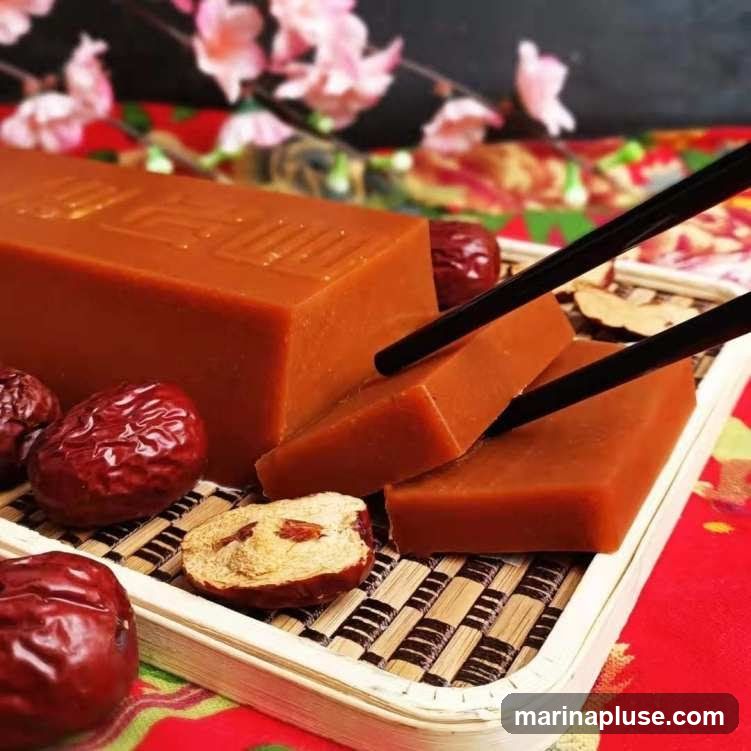 A rectangular block of reddish-brown cake, possibly jujube cake, is presented on a woven bamboo tray. Several whole dried jujubes and one halved dried jujube are scattered around the cake. Black chopsticks are used to pick up two slices of the cake. The background features a blurred image of pink cherry blossoms and a red patterned cloth.