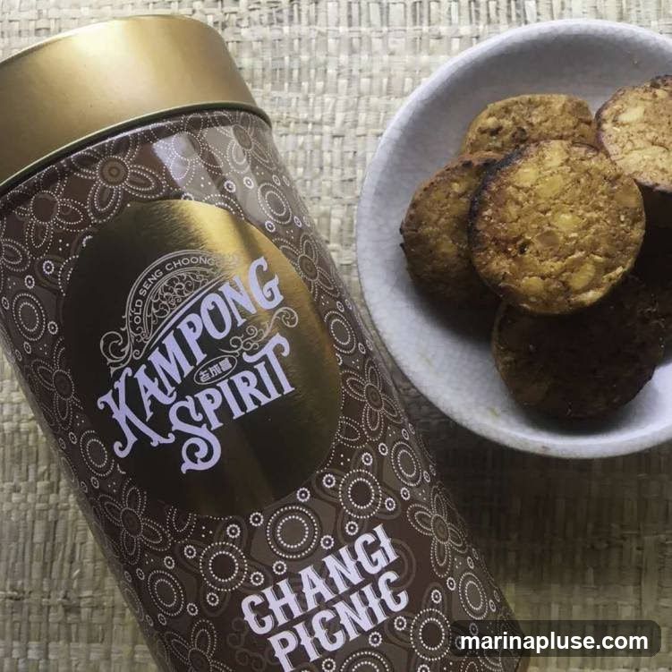 A close-up shot of a decorative tin labeled "Kampong Spirit" and "Changi Picnic" next to a small white bowl filled with round, golden-brown cookies. The tin has a brown background with a repeating pattern of white floral and circular motifs. The label features the words "Kampong Spirit" in a stylized font, with smaller text above reading "OLD SENG CHOONG". The cookies appear to be dense and possibly contain nuts or seeds.