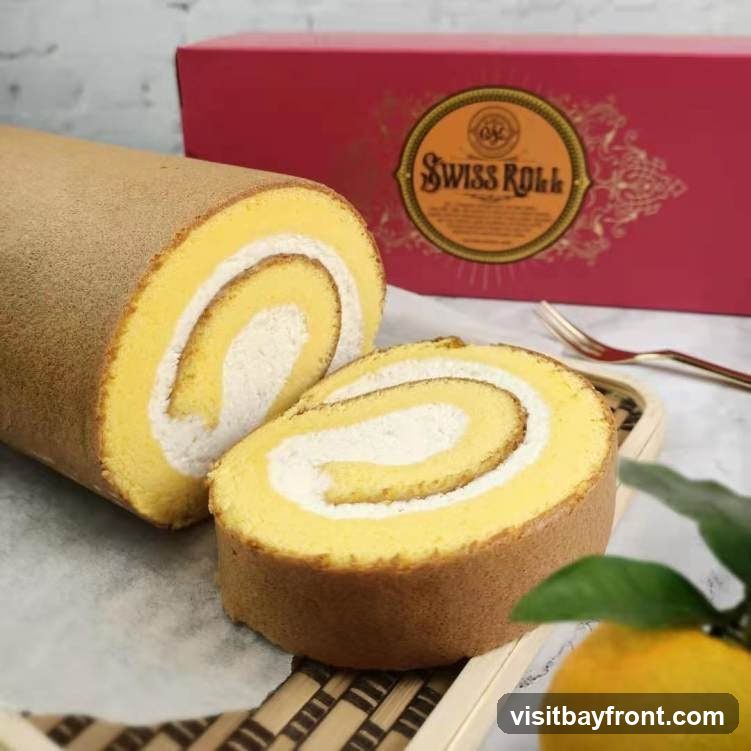 A close-up shot of a Swiss roll cake, with one whole roll and several slices on a serving platter. The cake has a light yellow sponge and a white cream filling. In the background, a red box with a circular logo that reads "Swiss Roll" is visible. A fork and a sprig of leaves with a citrus fruit are also in the frame.