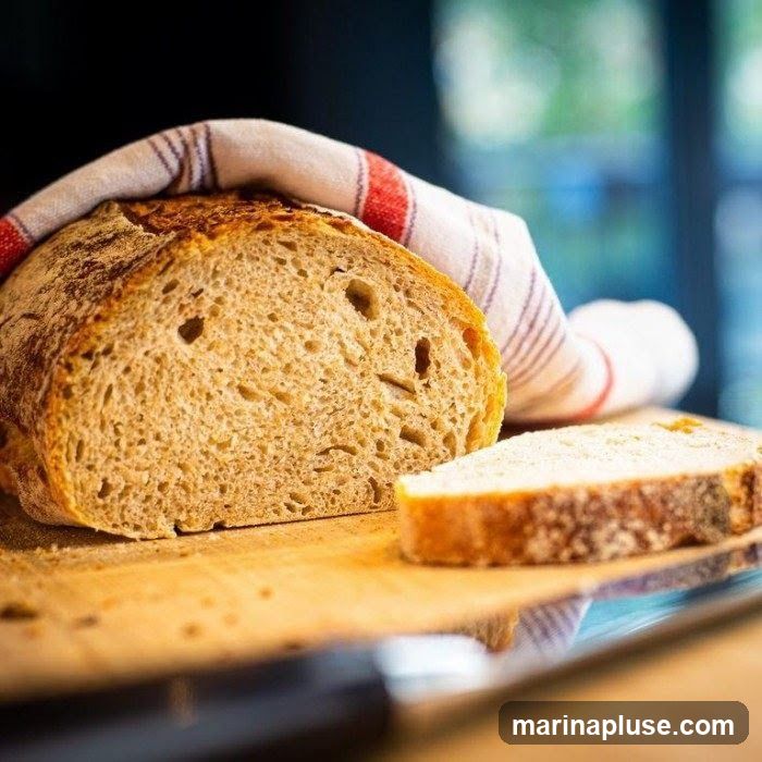 A close-up shot of a rustic loaf of bread, partially sliced, resting on a wooden cutting board. A striped kitchen towel is draped over the top of the loaf. The bread has a golden-brown crust and an airy crumb.