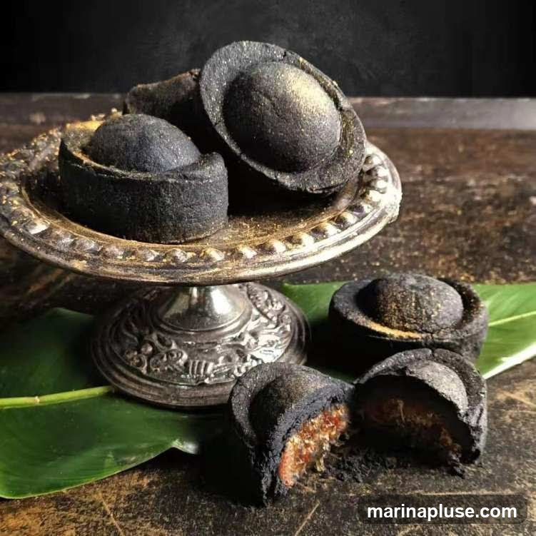 A close-up shot of several dark, round pastries dusted with gold powder. Some are arranged on an ornate silver pedestal dish, while others are placed on green leaves on a textured surface. One pastry is cut in half, revealing a reddish-brown filling.