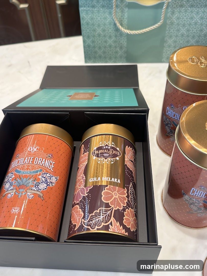 A black gift box is open, revealing two decorative tins of cookies. The tin on the left is orange with a "Chocolate Orange Sea Salt" label and a pineapple motif. The tin on the right is brown with a "Gula Melaka" label and floral patterns. A teal gift bag with a patterned design and rope handles is visible in the background. Other cookie tins with similar designs are also present.