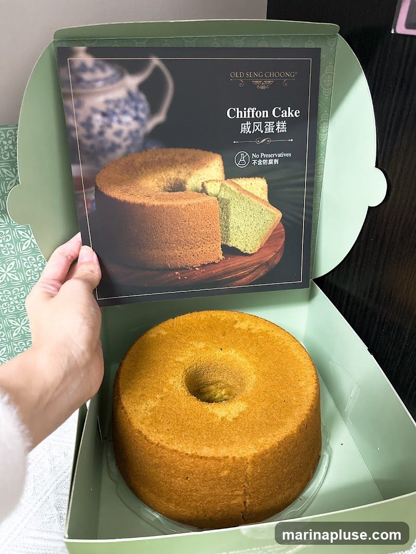 A hand holds a product insert over a chiffon cake in a light green box. The insert features an image of two chiffon cakes, one whole and one sliced, with text that reads "OLD SENG CHOONG Chiffon Cake" and "No Preservatives". The cake in the box appears to be a plain chiffon cake.