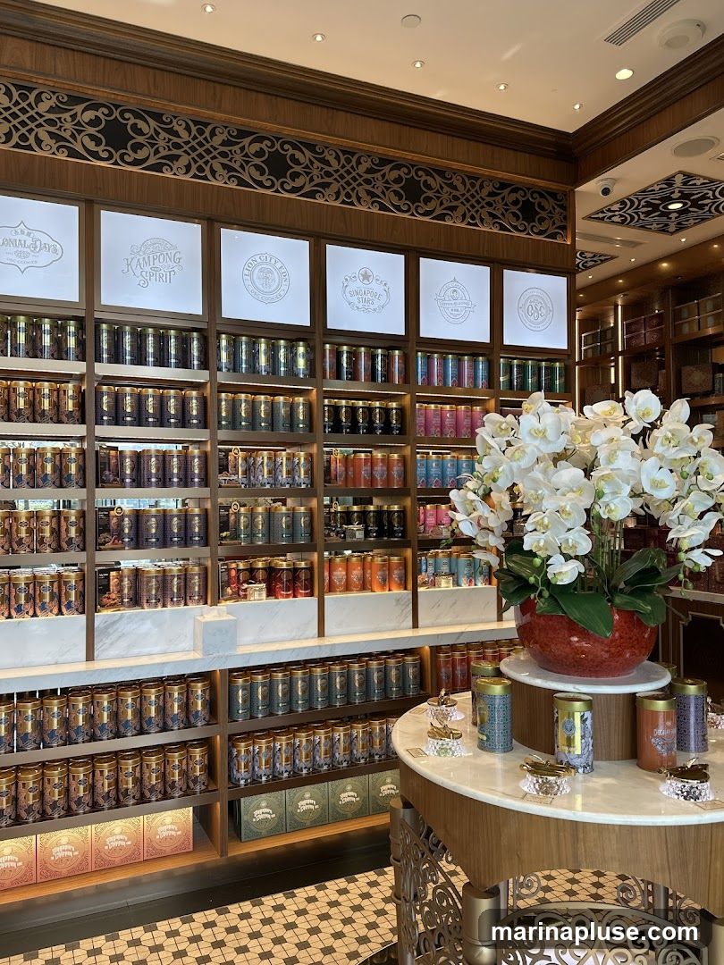 A well-lit interior of a shop filled with shelves of tea tins. The shelves are organized by color and design, with various sizes of tins. Above the shelves, there are framed logos or designs. A round display table in the foreground holds smaller tins and decorative items, topped with a large arrangement of white orchids in a red vase. The floor has a patterned tile design.