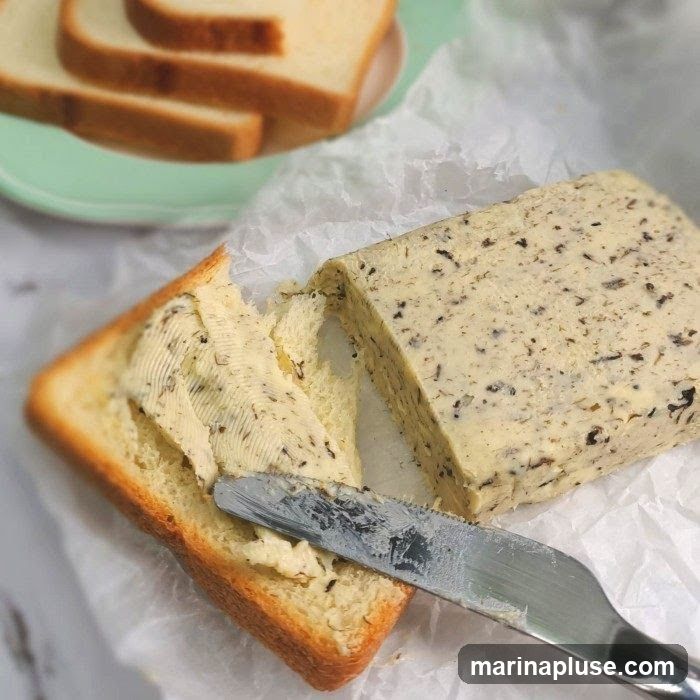A slice of toast is being buttered with a knife. A block of herb butter sits on parchment paper next to the toast. Several slices of toast are on a green plate in the background.