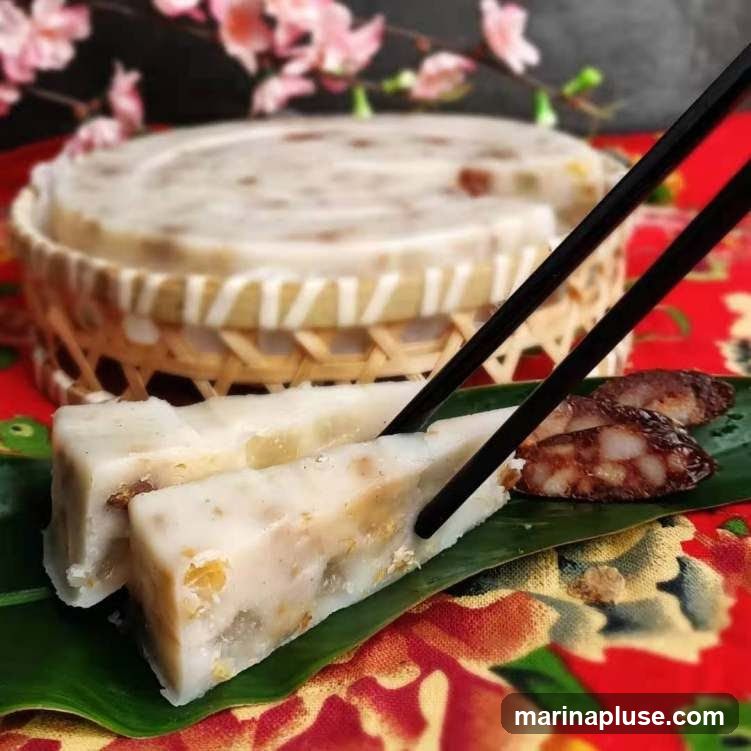 Two pieces of turnip cake are being picked up by chopsticks from a banana leaf. In the background, a whole turnip cake is in a woven basket. Slices of Chinese sausage are on the banana leaf next to the turnip cake. The table is covered with a red cloth with a floral pattern. Pink cherry blossoms are blurred in the upper left corner.
